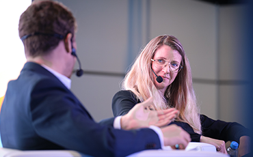 Two speakers engage the audience on stage at a bustling show floor, sharing insights and expertise.